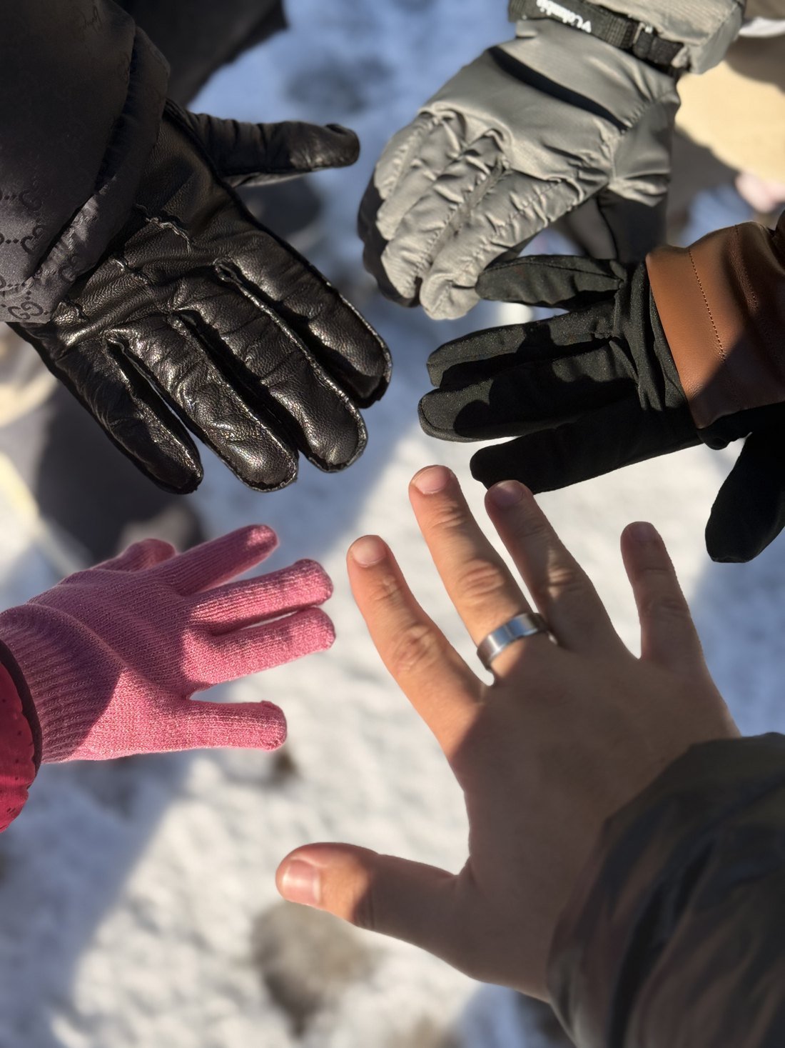 Cinco mãos da família, com luvas de tamanhos diferentes e uma aliança em primeiro plano, sobre a neve